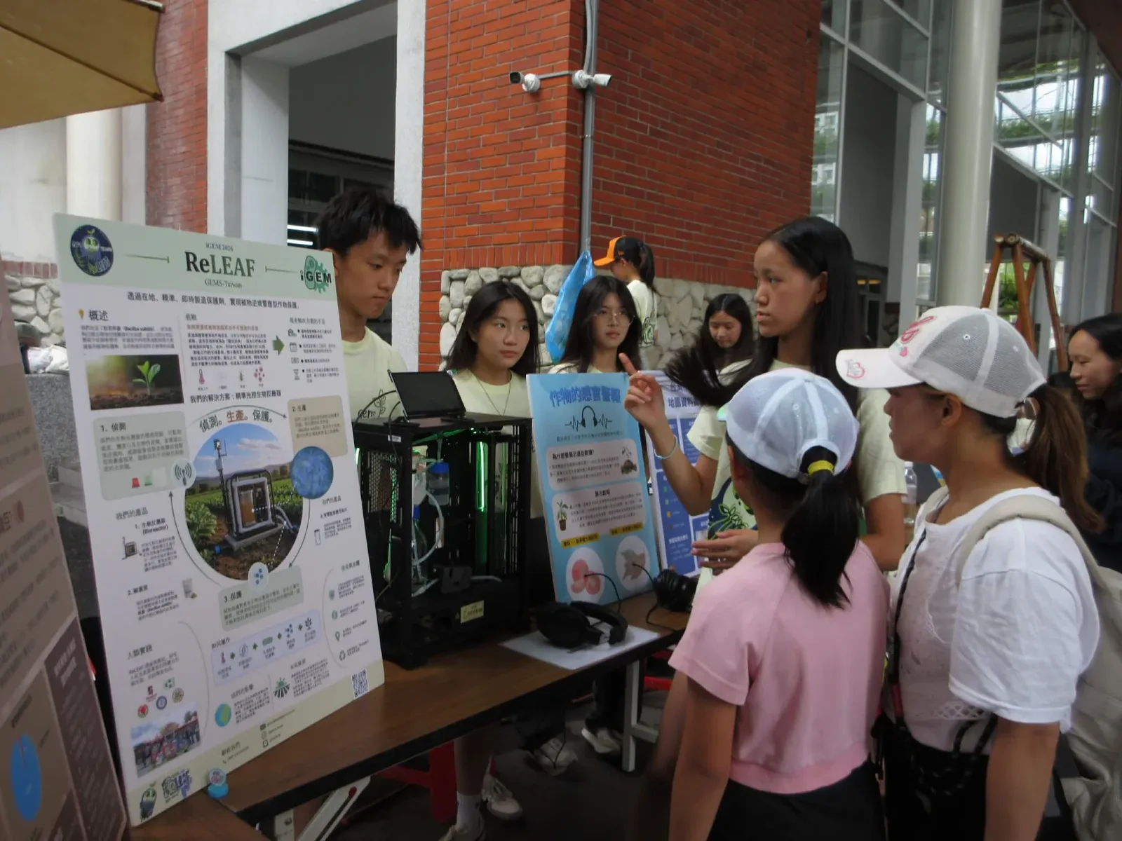 The ReLEAF booth at an outdoor market. A team member points at a blue poster headed 作物的感官警報 while visitors, two of them children in caps, look on. The bioreactor prototype sits on the table beside a pair of headphones, next to the project poster.