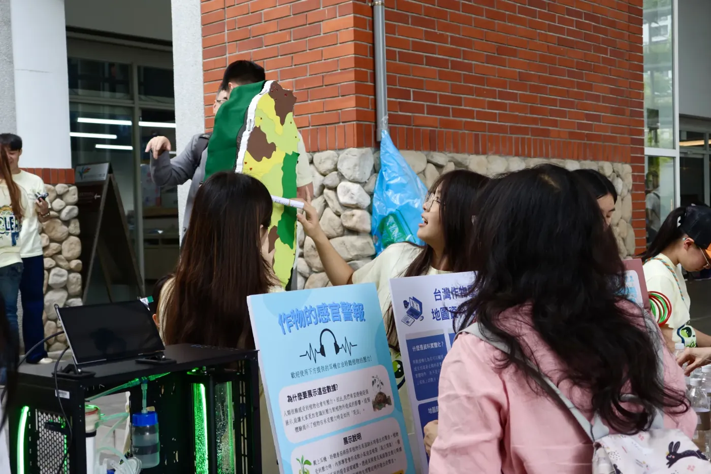 A team member holds the styrofoam map of Taiwan up against a brick wall while visitors crowd in front of it. The island is covered in dark green, yellow-green and brown paper. In the foreground a laptop and the blue sound-exhibit board stand on the table.