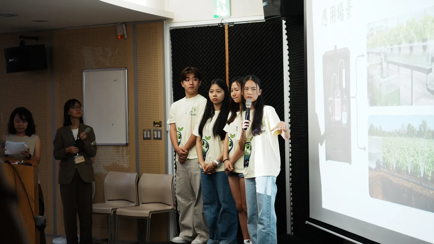Four ReLEAF students stand beside a projection screen headed with the Chinese words for application scenarios, which shows the bioreactor linked by arrows to a hydroponic greenhouse and to a soil-grown field. One student speaks into a microphone.