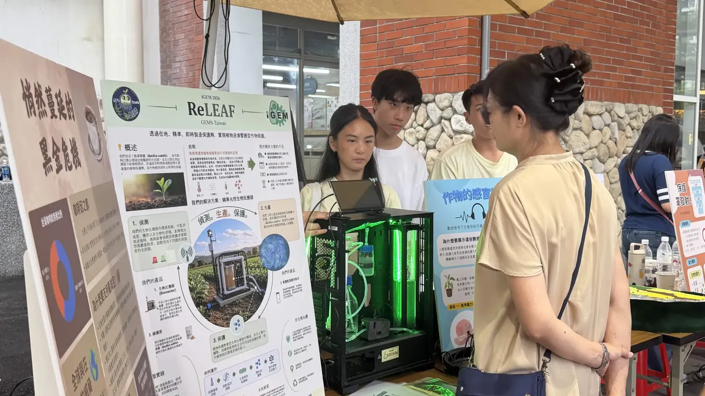 Visitors talk with students beside the green-lit ReLEAF bioreactor and a team poster at an outdoor forum booth.