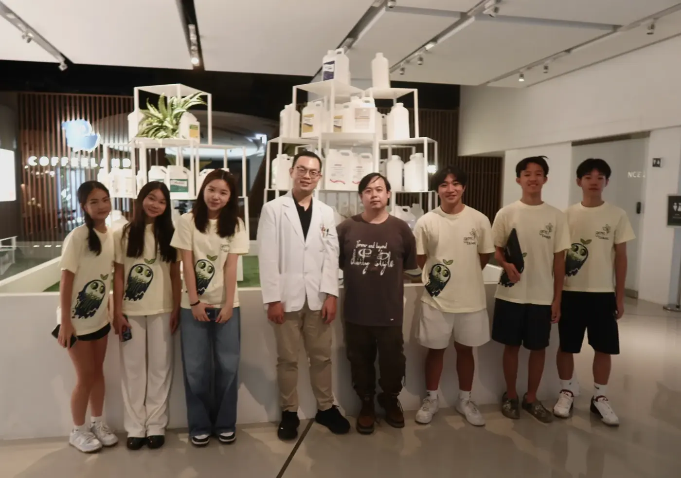 Eight team members stand with two hosts in a bright showroom with potted plants on white shelves.