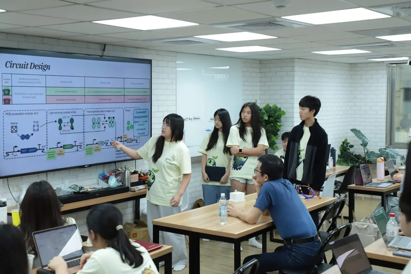 A student presents a circuit design slide to a seated adviser while the team stands around the screen.
