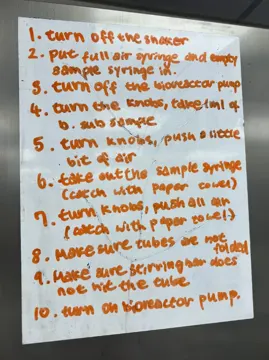 Whiteboard in orange marker listing steps 1 to 10 for taking a 1 ml B. subtilis sample from the bioreactor with syringes and knobs.
