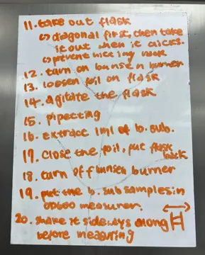 Whiteboard in orange marker listing steps 11 to 20 for sampling the flask next to a Bunsen burner and measuring OD600.