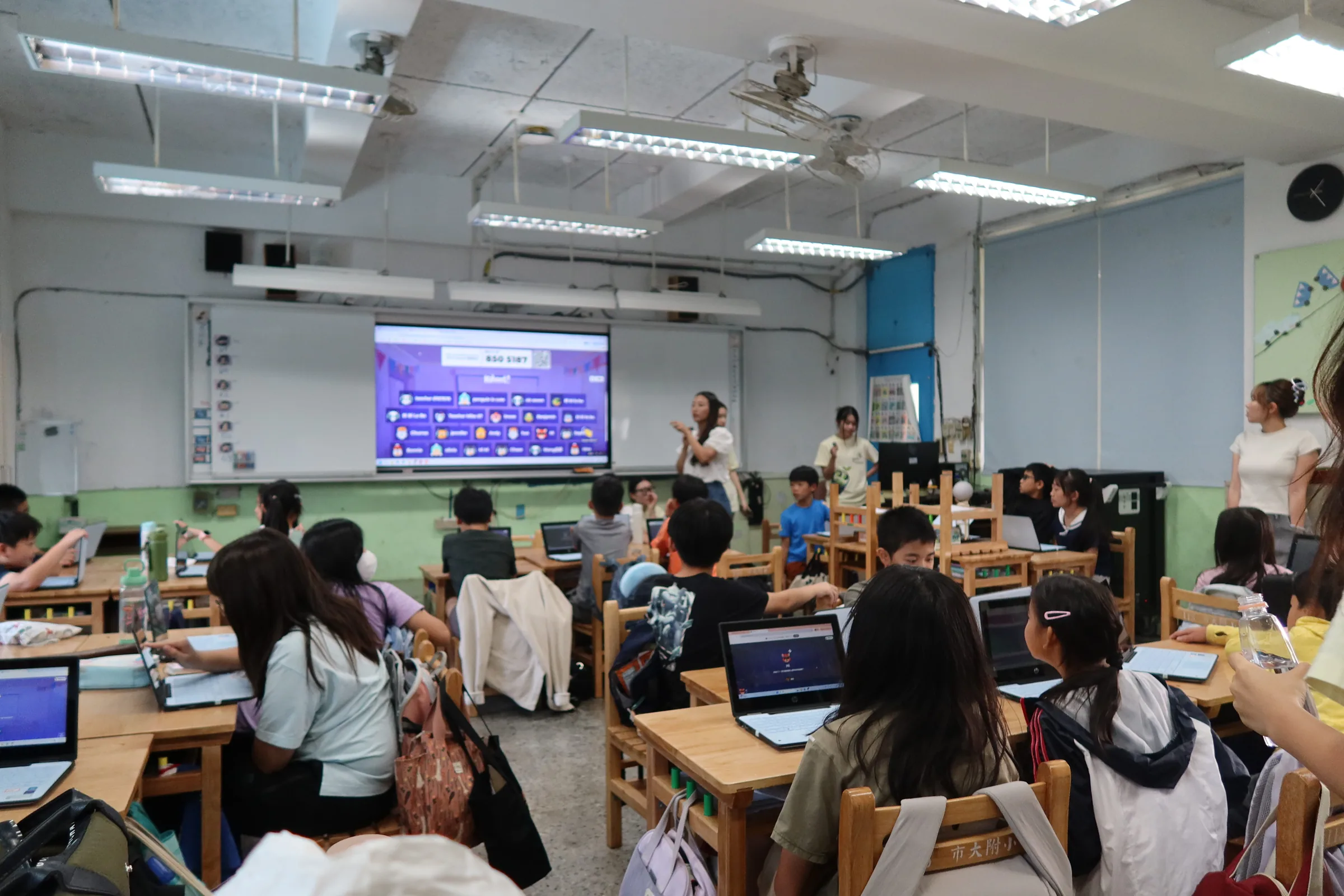 A classroom of elementary students at laptops facing a projected Kahoot quiz, with team members standing at the front.