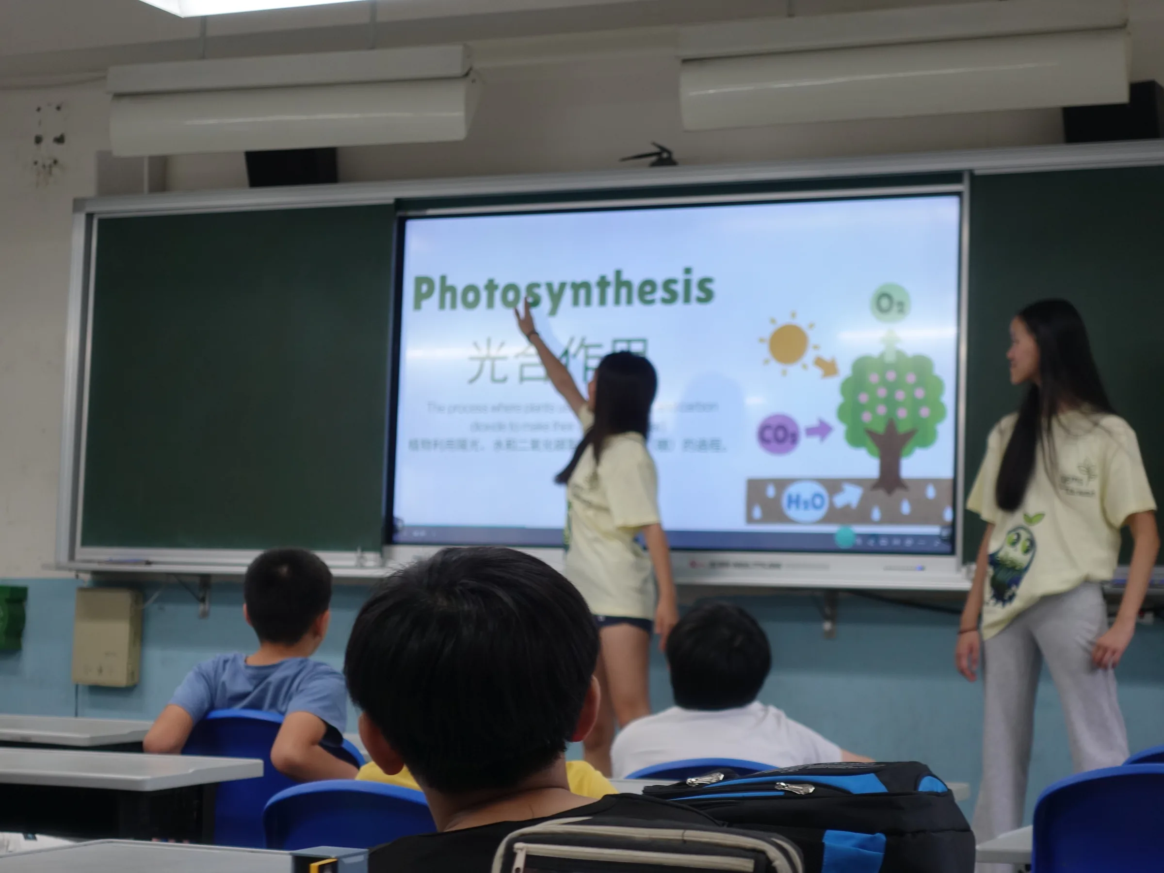 A team member pointing at a projected Photosynthesis slide while seated elementary students watch.