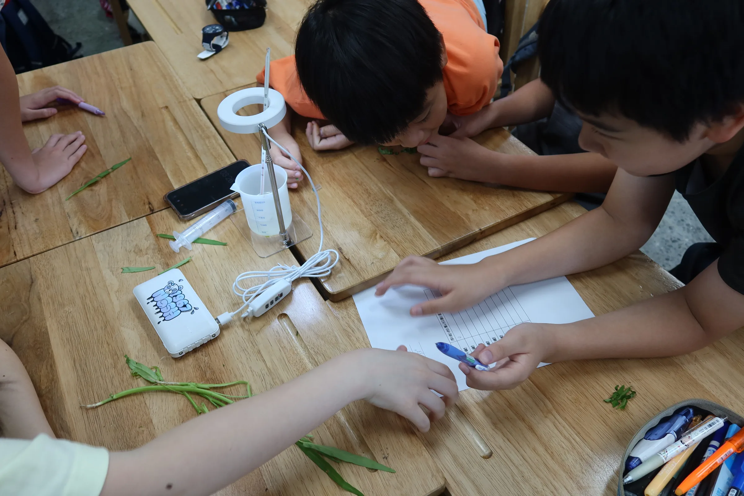 Students leaning over a wooden desk, writing on a worksheet beside a beaker under a small lamp.
