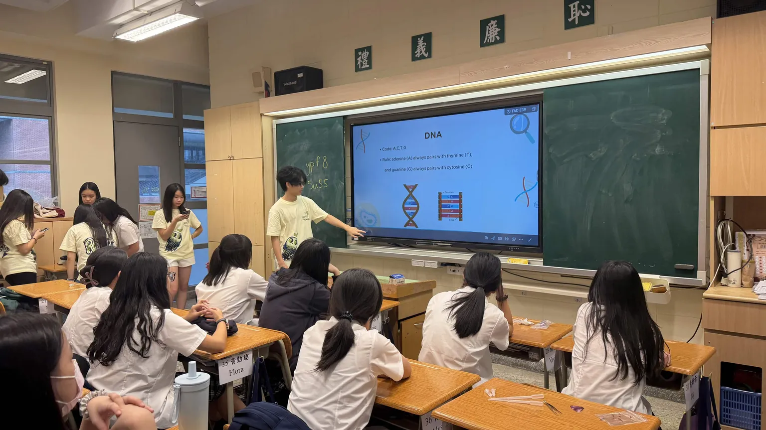 Team members teaching beside a projected slide titled DNA with a double-helix diagram, students seated at their desks.
