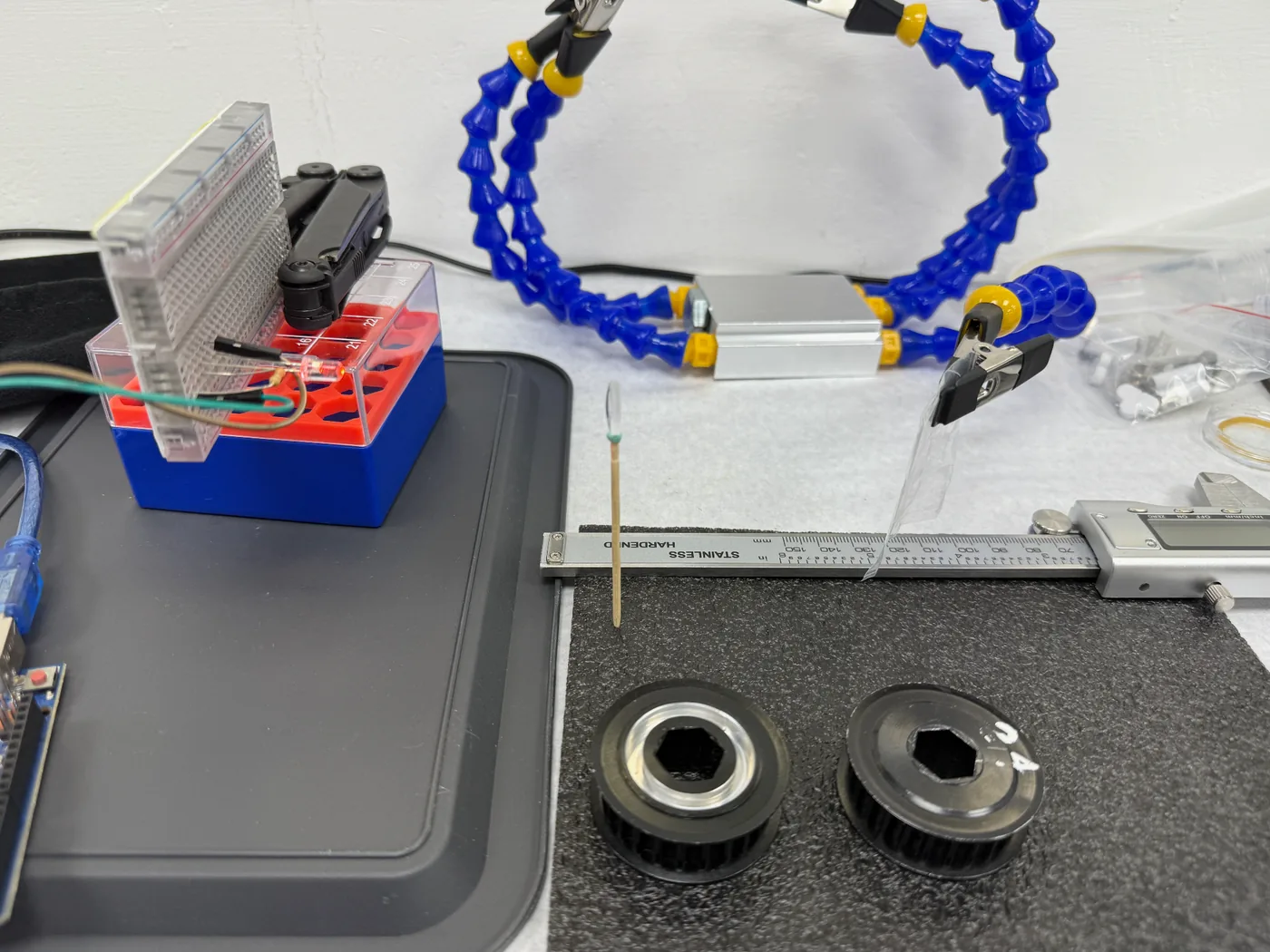 A bench alignment rig: blue magnetic helping-hands arms holding a beam splitter in an aluminium housing and a cuvette in line, a steel rule and a digital caliper laid under them, with a breadboard and the LED on the left.