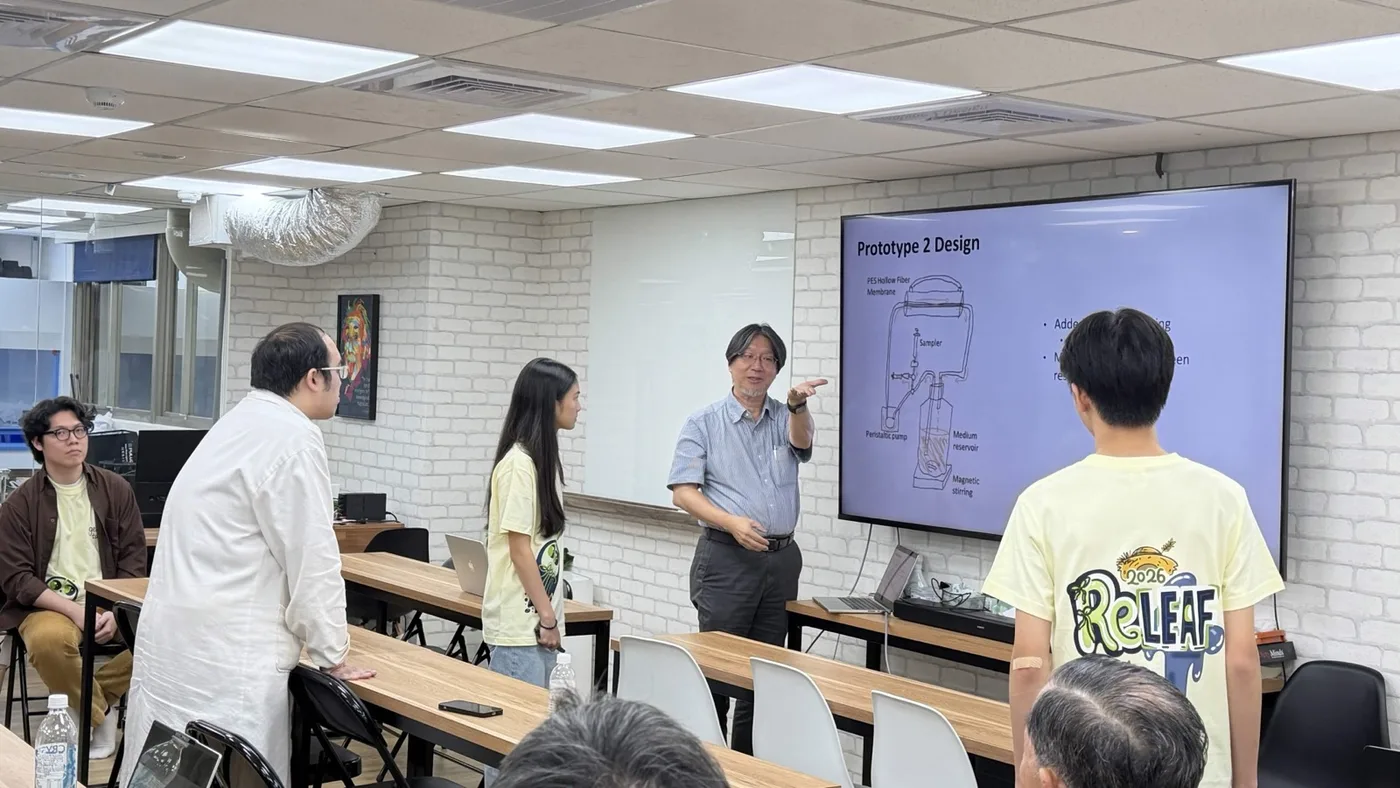 A projected schematic of prototype II under review at a table, showing membrane, sampler, peristaltic pump, medium reservoir and magnetic stirring, with team members discussing it.
