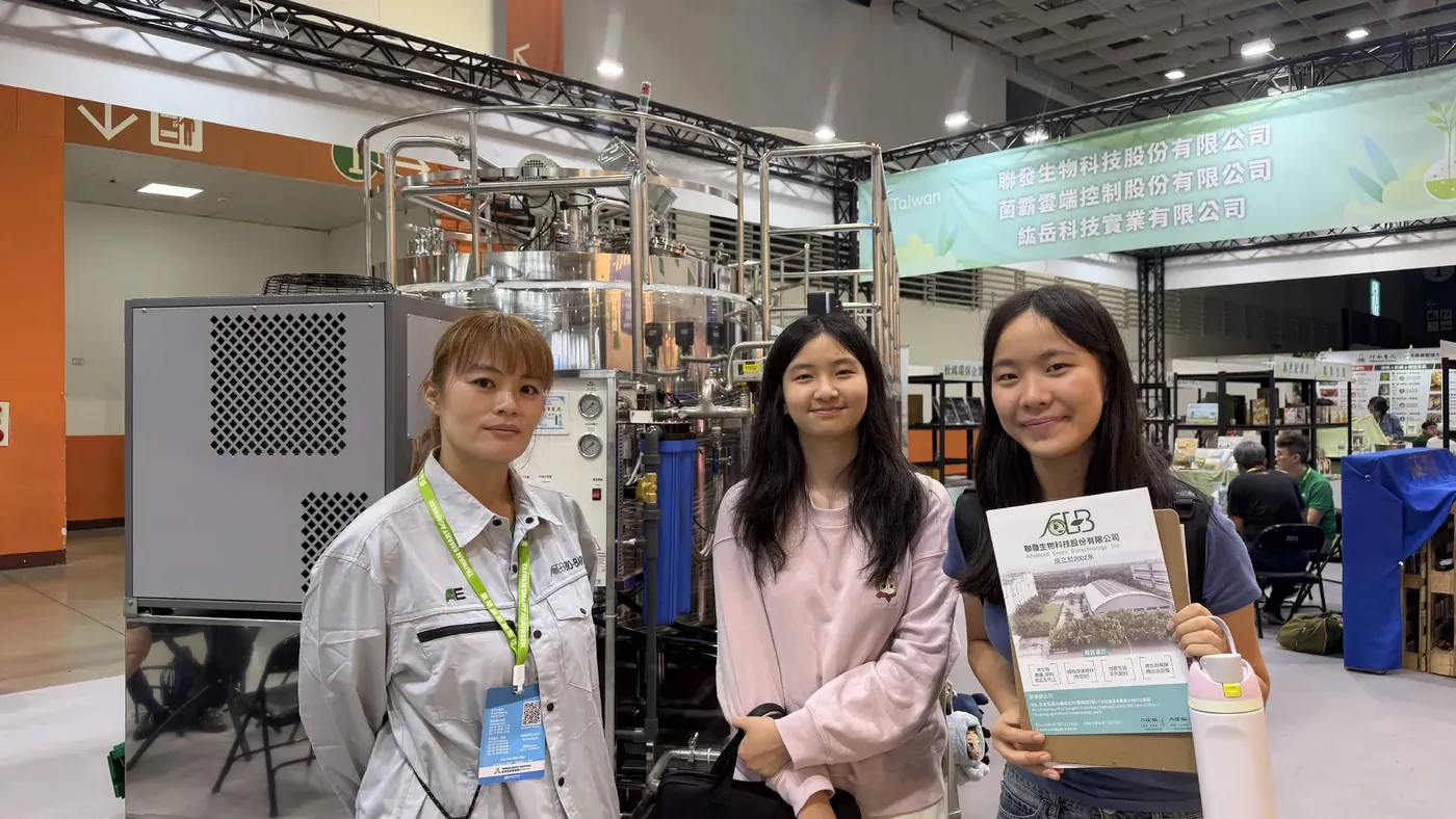Two ReLEAF students standing beside an exhibitor in a work jacket at a trade show booth, one holding a company brochure, with a stainless steel fermentation system behind them.
