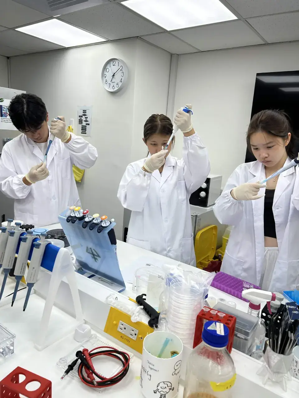 Three team members in lab coats pipetting at the bench, with racks and tip boxes in front of them.