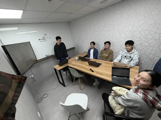 Five students around a wooden table with laptops in a small meeting room, one standing beside a whiteboard.