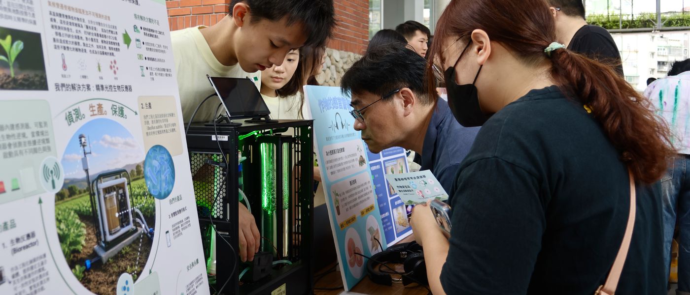 At the 5 September forum booth, a ReLEAF student leans over the black bioreactor cabinet, its tubes lit green, while two visitors bend in to look; the team&rsquo;s poster stands to the left.