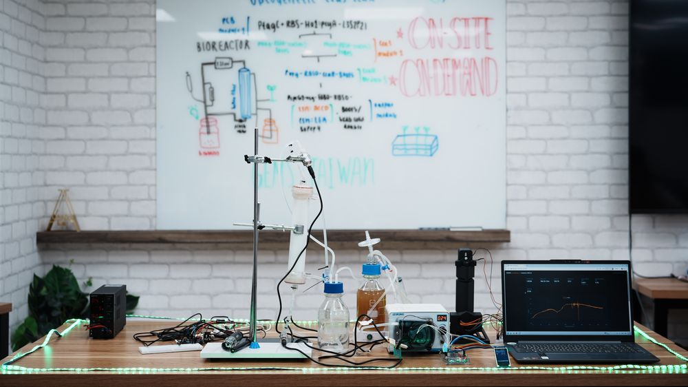 The ReLEAF system running on one bench: a hollow-fiber column on a stand, culture and medium bottles, a peristaltic pump, the in-line photometer and a laptop showing a live growth curve, under a whiteboard sketch of the circuit and the bioreactor loop.