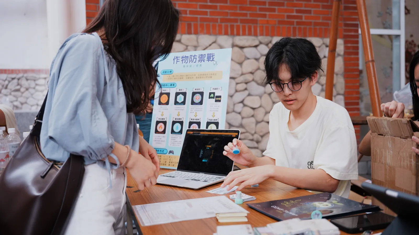 A student guides a visitor through a game on a laptop.