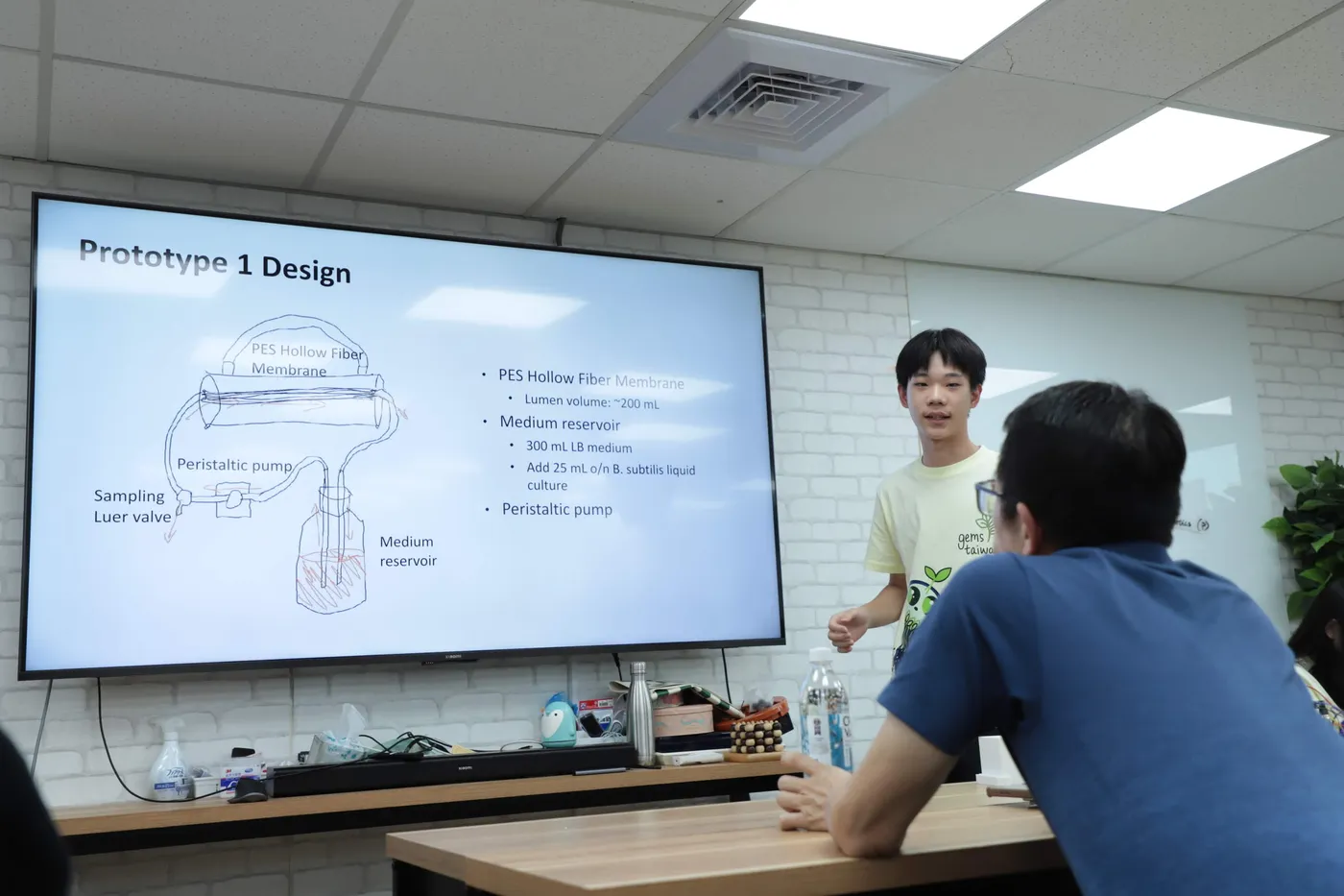A student presenting a slide headed Prototype 1 Design, showing a hand-drawn PES hollow fibre membrane, peristaltic pump and medium reservoir, to a seated listener in a meeting room.
