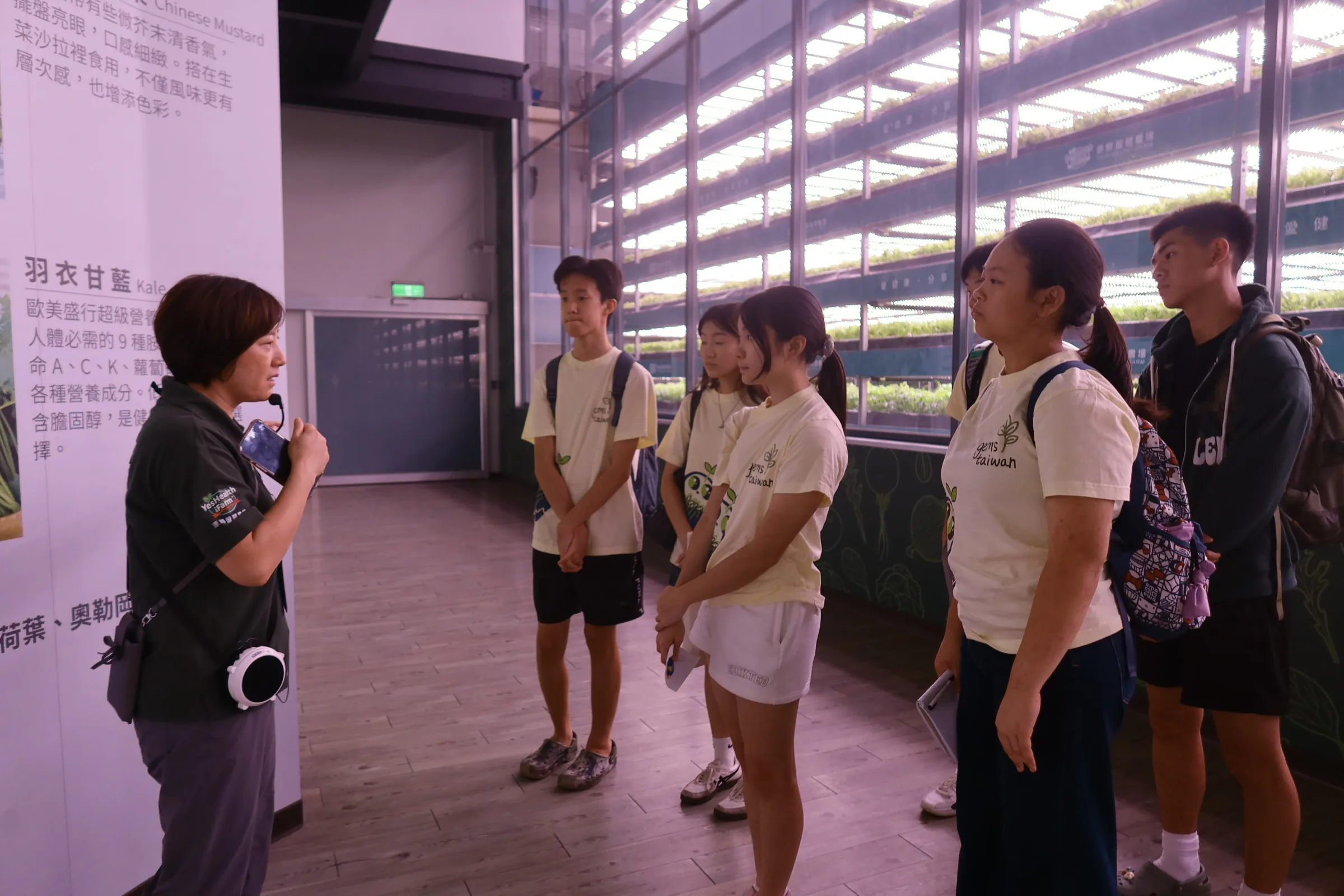 A guide with a portable speaker talking to five students beside tall racks of lit growing shelves.