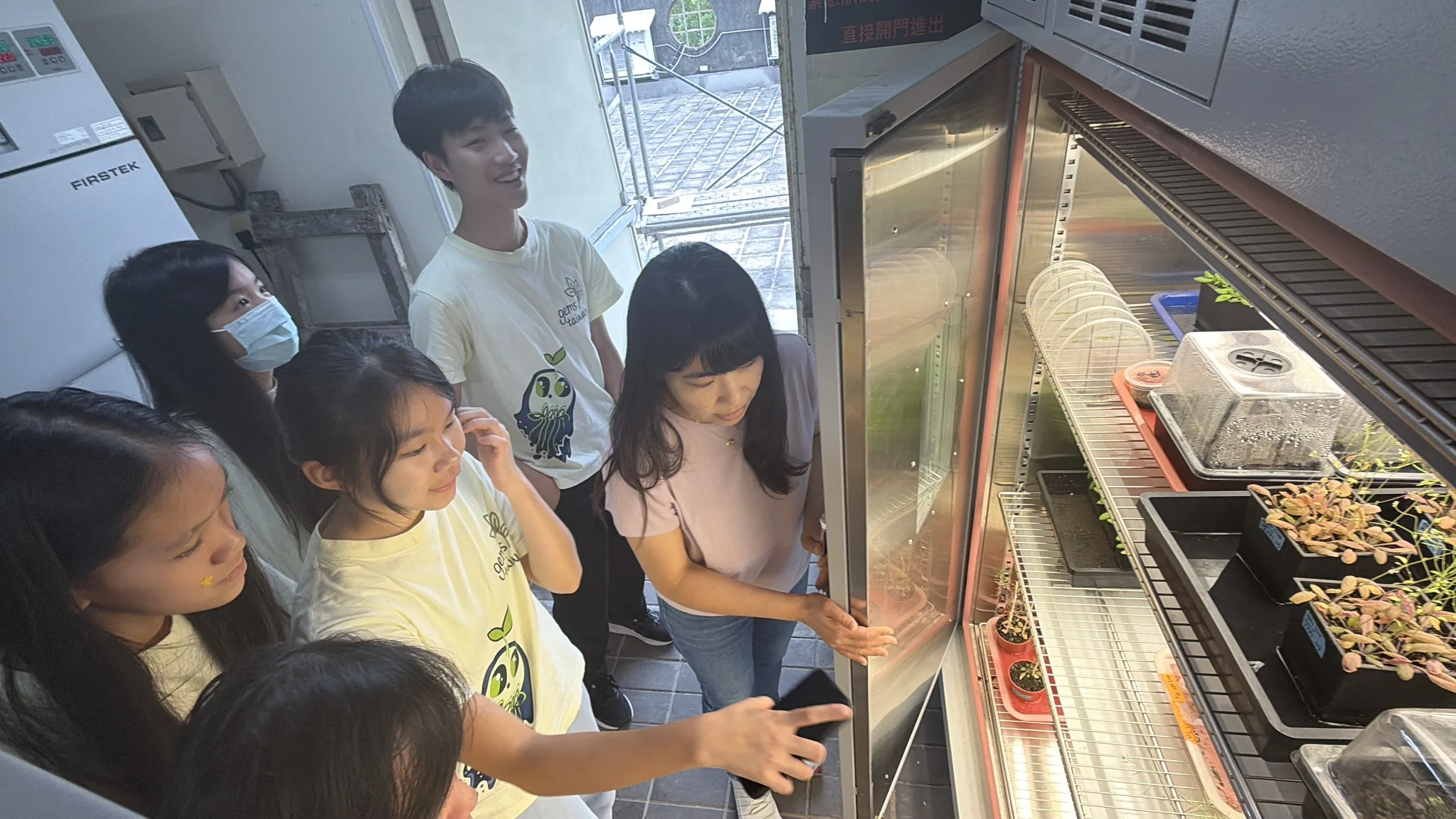 Students with Prof. Cheng looking into an open growth chamber holding trays of plants.