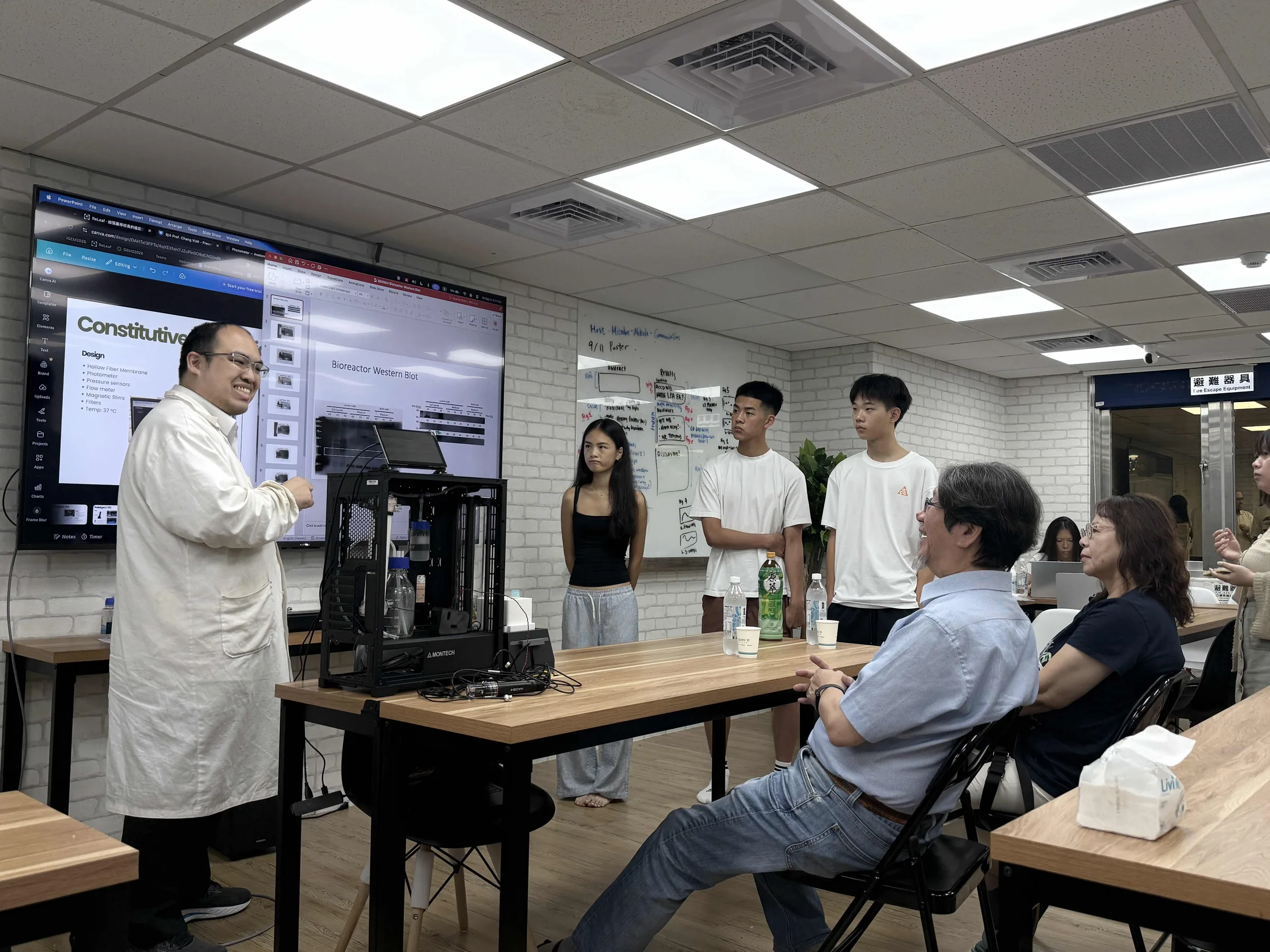 A presenter in a white lab coat beside the bioreactor frame on a table, with Prof. Chang seated in front and students standing behind.
