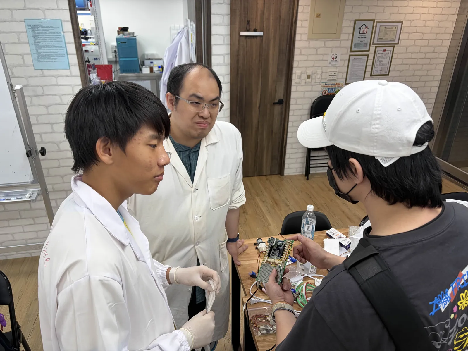 Teacher He in a white lab coat with three students around a circuit board and wiring on the lab bench.