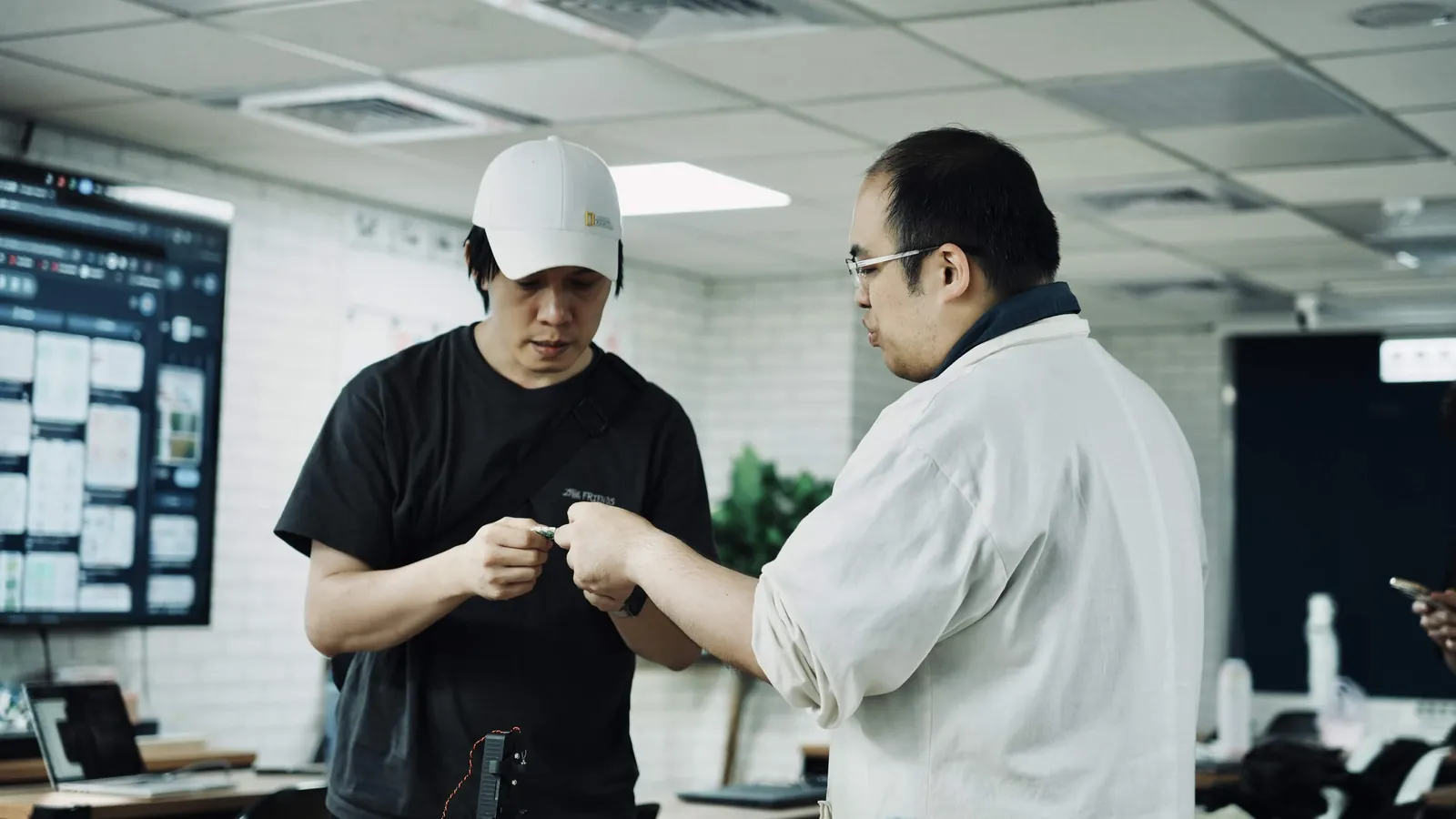 A student in a white cap and Teacher He, in glasses, looking down together at a small part one of them is holding, in a room with a dashboard on a wall screen.