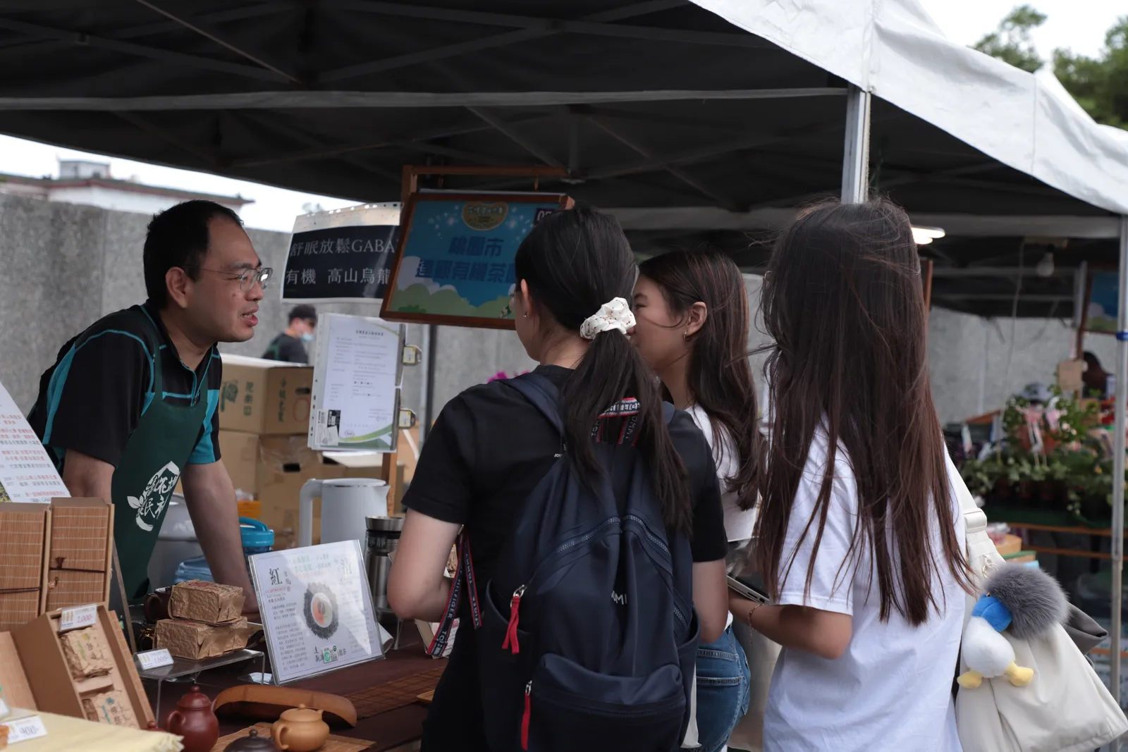 A grower in a green apron explaining his produce to three students at his stall.