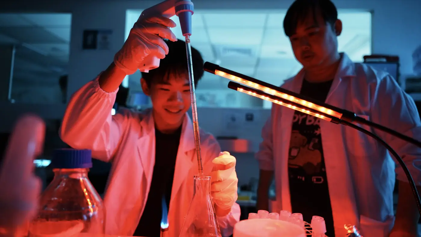 A team member pipetting into a flask under a red safelight while a second person watches over their shoulder.