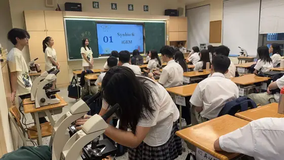 A high school classroom with students in uniform at desks, several microscopes on a bench in the foreground, and a ReLEAF member presenting a slide headed Synbio and iGEM at the front.