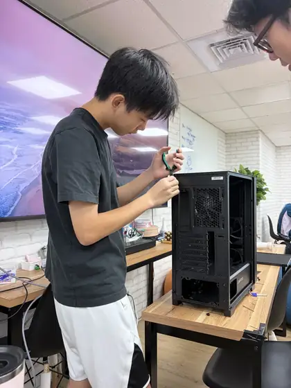 A student standing at a table using a screwdriver on the frame of an empty black computer tower case, with another student watching from the right.
