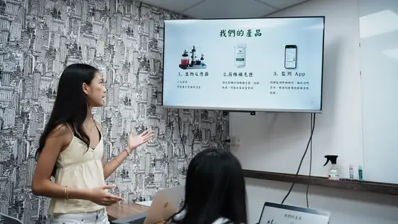 A team member rehearsing in front of a wall screen showing a slide headed 我們的產品, our products, with phone interface mockups, while a seated listener takes notes.