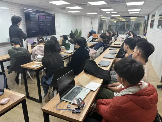 A long room of students at wooden benches on the first day of the season, with a laptop and a wired-up microcontroller board on the nearest desk in the foreground.