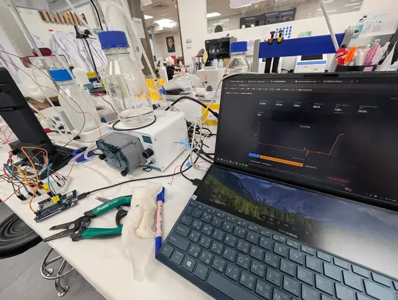 The inline photometer plumbed into the bioreactor loop on a bench, with a laptop beside it showing a rising orange optical density trace.
