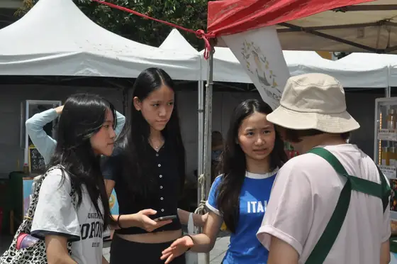 Three students standing in a market aisle talking with a grower in a wide-brimmed bucket hat, one holding a phone to record notes.