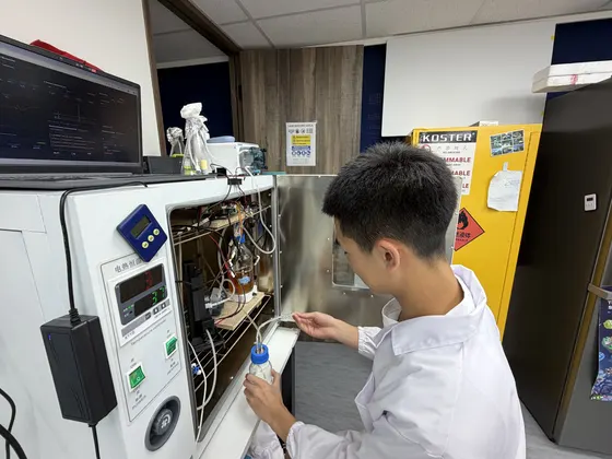 A student in a lab coat crouched at the open bioreactor cabinet at night, taking a sample from the loop.