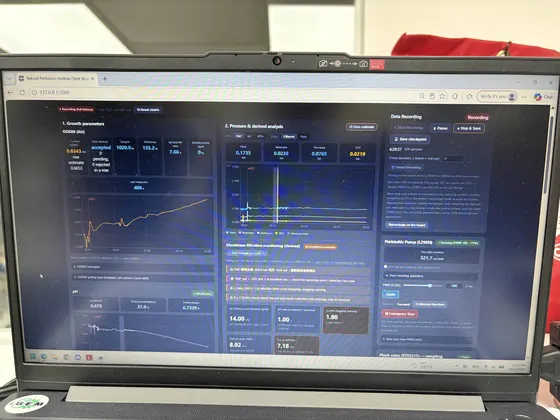 A laptop on a bench showing the dashboard: a growth curve climbing on a dark blue panel beside live pressure and pH readings.
