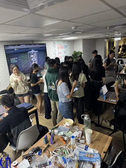 The work room full of students standing and sitting at every desk, the nearest table covered in parts, cables and food.