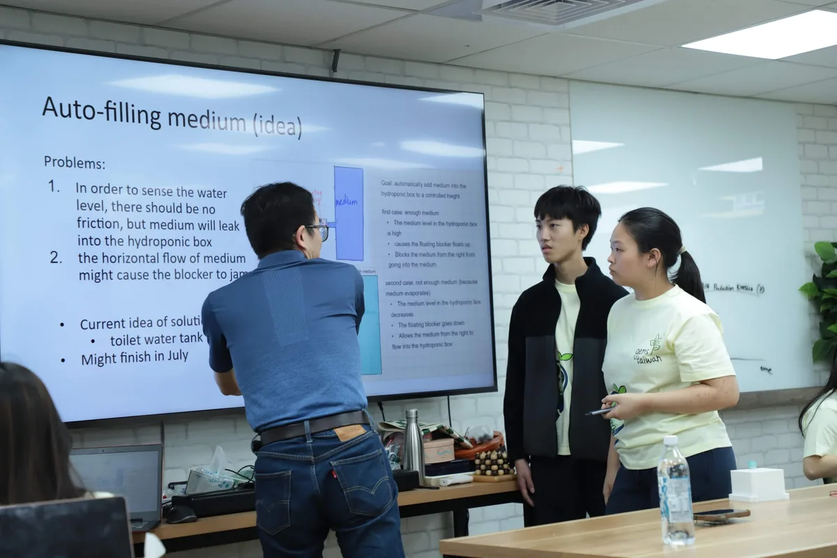 Prof. Chen standing beside a wall display showing the team's own slide about auto-filling medium, two students standing with him.