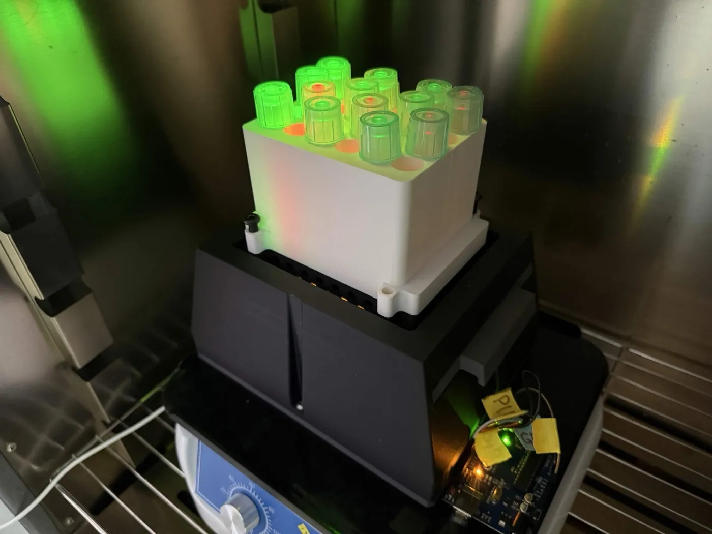 The light-programmable array running inside an incubator: a white block of capped culture tubes glowing green and red from the LEDs beneath, mounted on a black base on a shaker.