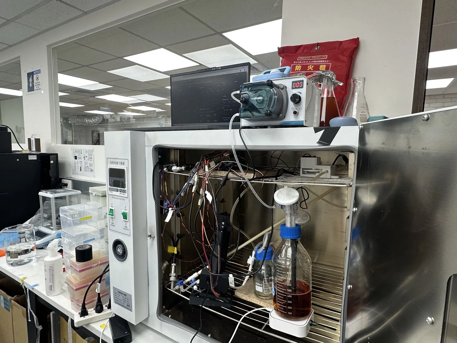 The bioreactor inside an incubator with its door open: a stirred bottle of amber culture, tubing, the black in-line photometer and wiring, with a peristaltic pump and a laptop showing the dashboard on top.