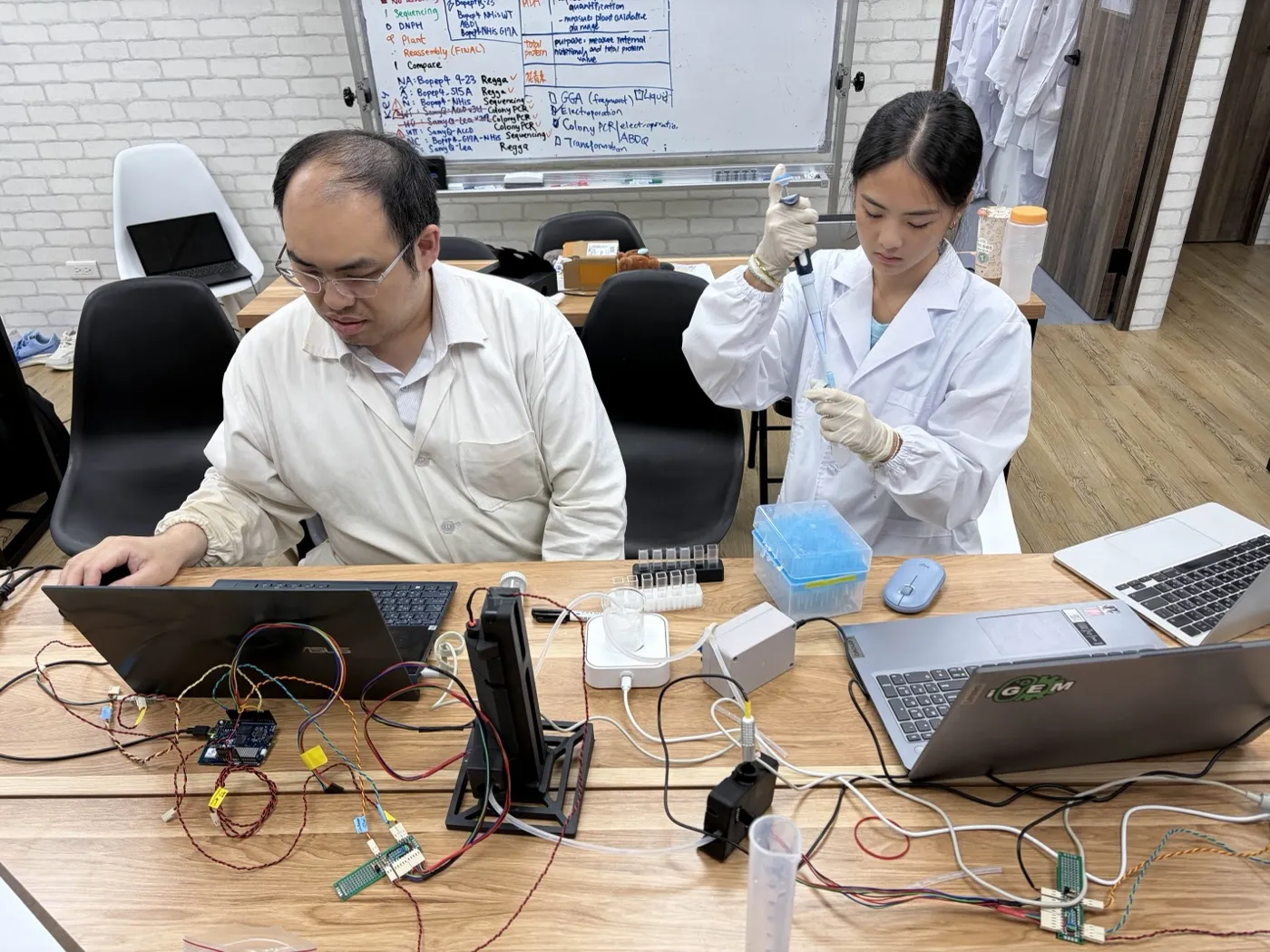 A student in a lab coat pipettes a sample into a cuvette beside the black in-line photometer and its stirred beaker, while an instructor logs the readings on a laptop.