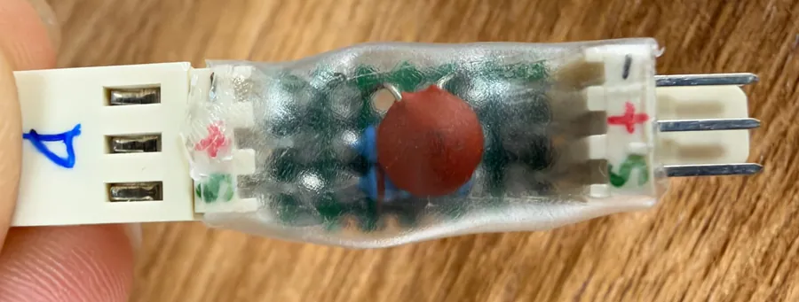 The adapter board sealed in clear hot glue, seen from its component side: a white three-pin socket on the left marked 4 in blue, a three-pin male header on the right, both with their pins hand-marked minus, plus and S, and a round red capacitor over a blue resistor in the middle