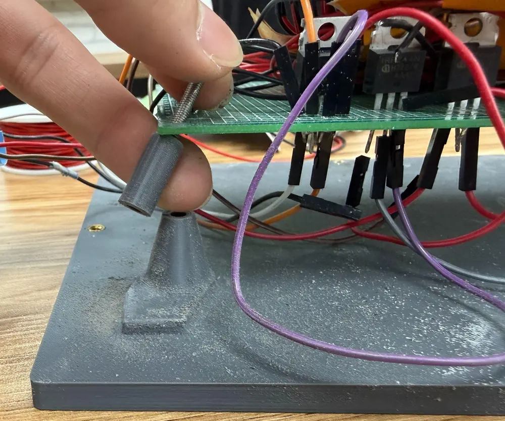 A grey printed bottom plate with a cone-shaped base; fingers hold the snapped-off cylindrical post just above it, a screw through its top, with the perf board, three MOSFETs and dust on the plate behind.