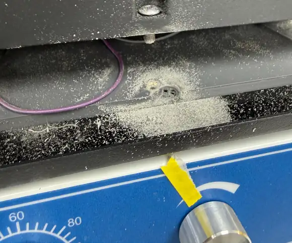 Close-up of the LPA on a lab shaker: the lower edge of the black housing with a screw, the grey bottom plate beneath it with white debris spread around a torn hole, a black rubber mat, and the shaker's blue front panel with a dial marked 60 and 80 and a strip of yellow tape by its knob.