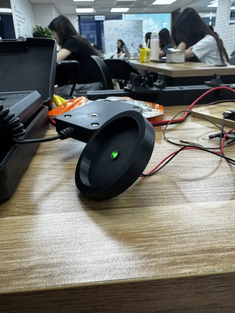 A round black printed fixture on a desk with one green LED lit at its centre, wired to the bench, the light meter's case behind it.