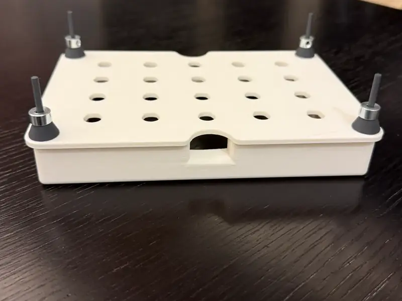 The first printed floating plate on a dark table: a white slab with a grid of holes over a closed hollow frame, a threaded pole with a washer stack at each corner.
