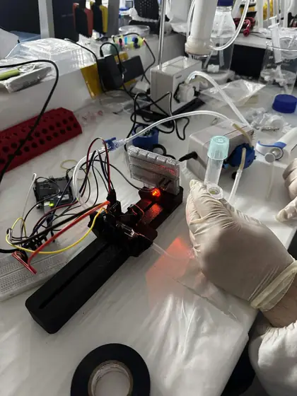 V1 on a lab bench: the black printed rail plumbed into tubing, its LED lit red, beside a gloved hand holding a sample tube
