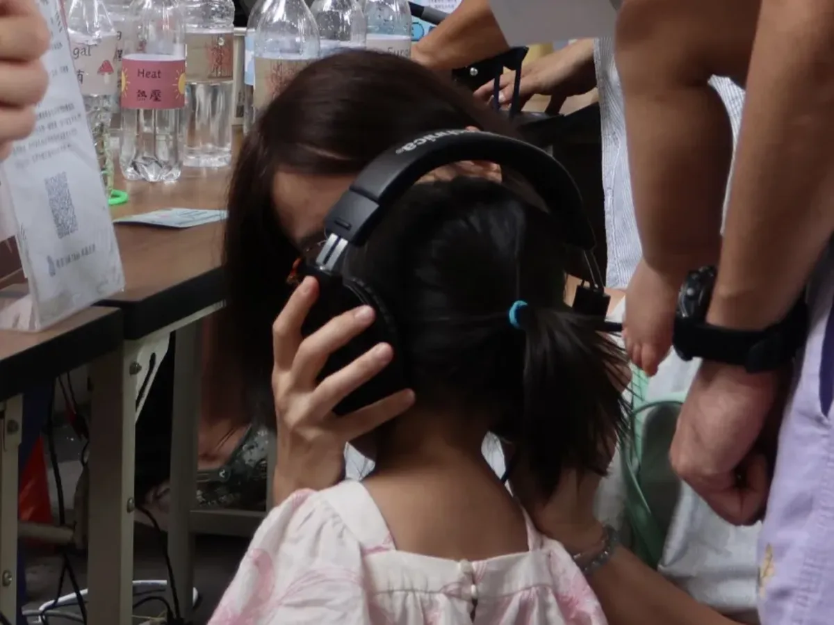 A young child at the booth, seen from behind, wearing large over-ear headphones held in place by an adult beside her. Behind them on the table stand labelled water bottles.