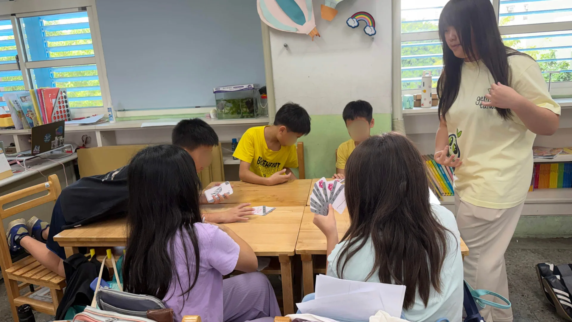 Elementary students sitting around a table holding cards, playing the ATCG base-pairing card game while a team member watches.