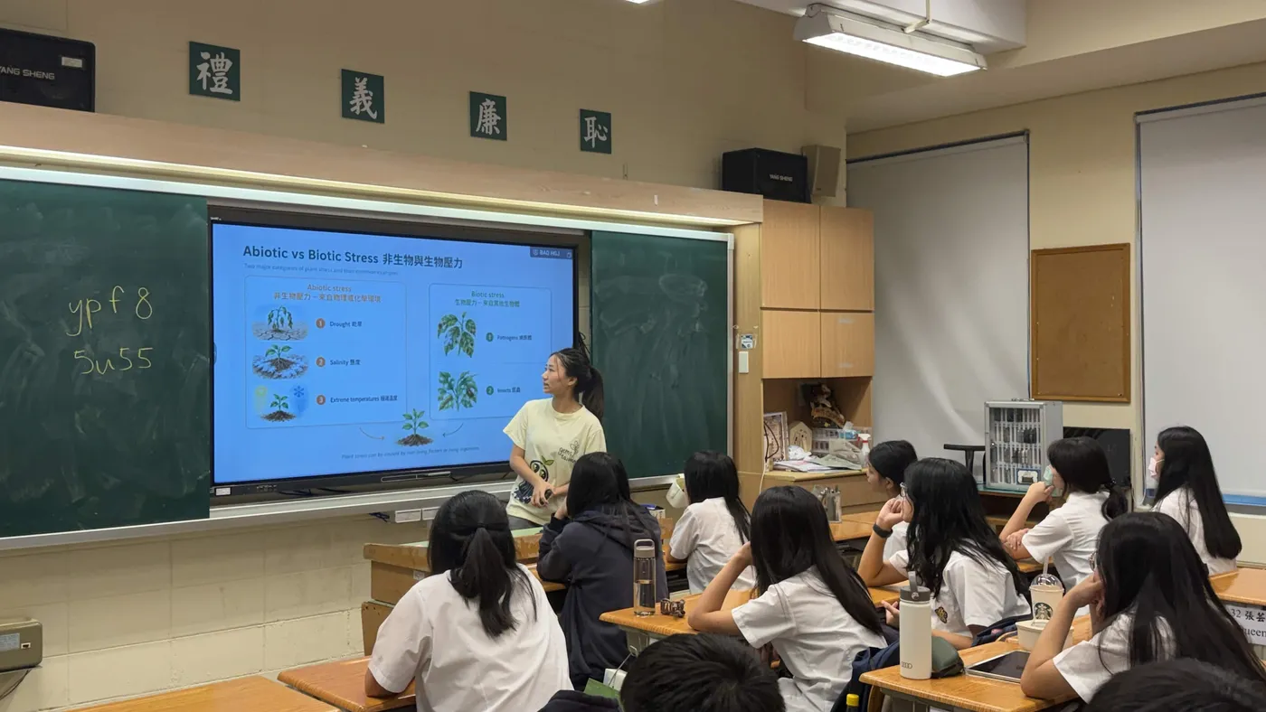 A ReLEAF team member teaching a high school class in front of a projected slide reading Abiotic vs Biotic Stress, with students seated at desks facing the board.